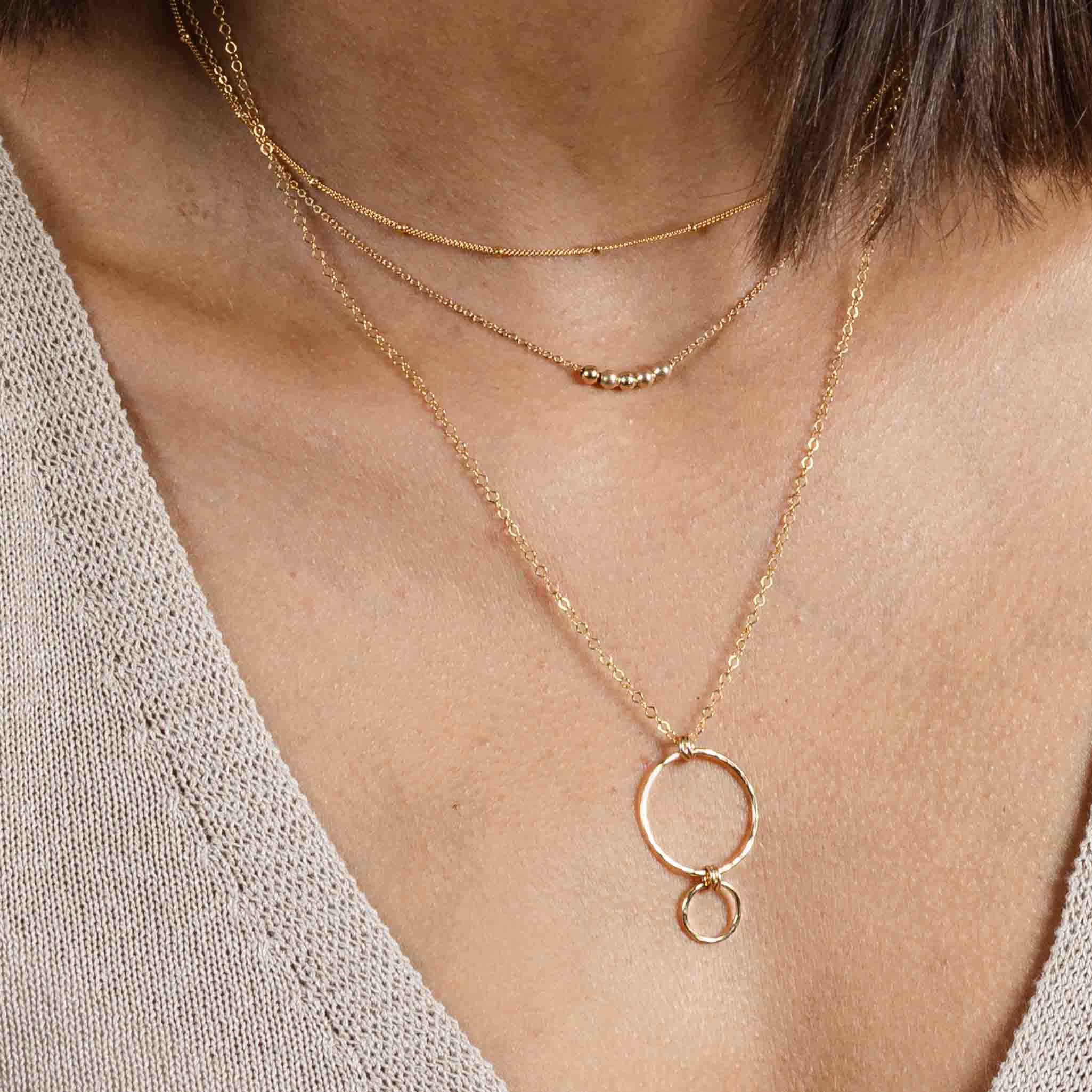 Am Hals des Models hängen drei Goldketten in verschiedenen Längen: eine ganz kurze Satellitenkette, eine feine Kette mit kleinen Goldperlen in der Mitte und eine längere Kette mit einem Anhänger, der aus gehämmerten Ringen besteht.