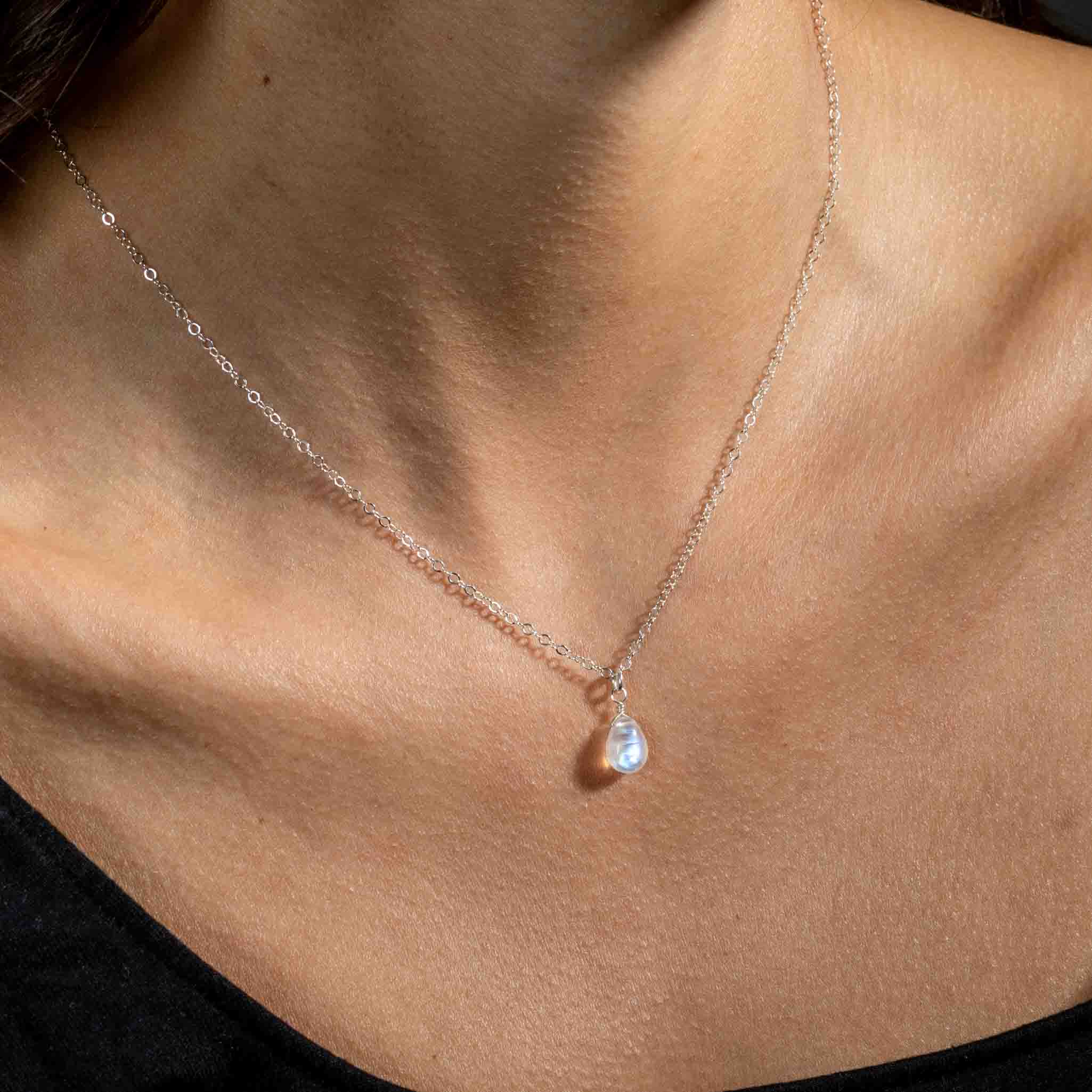 Eine feine Silberkette mit einem kleinen, bläulich schimmernden Mondstein-Anhänger in Tropfenform hängt am Hals des Models.