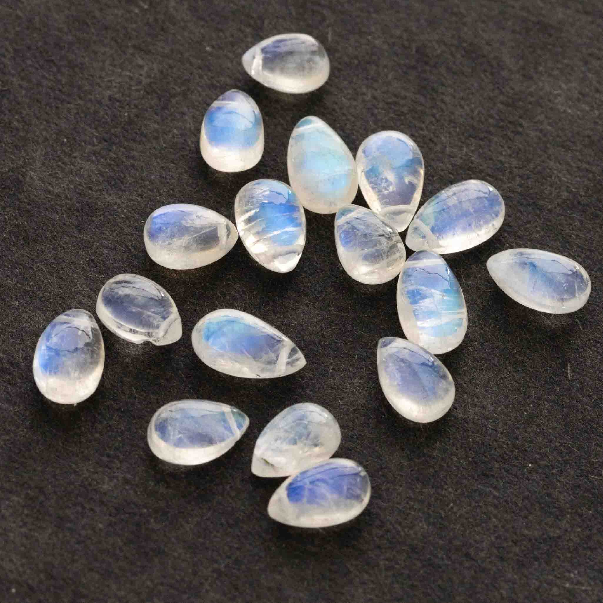Viele bläulich schimmernde Mondsteine in Tropfenform liegen auf schwarzem Hintergrund.