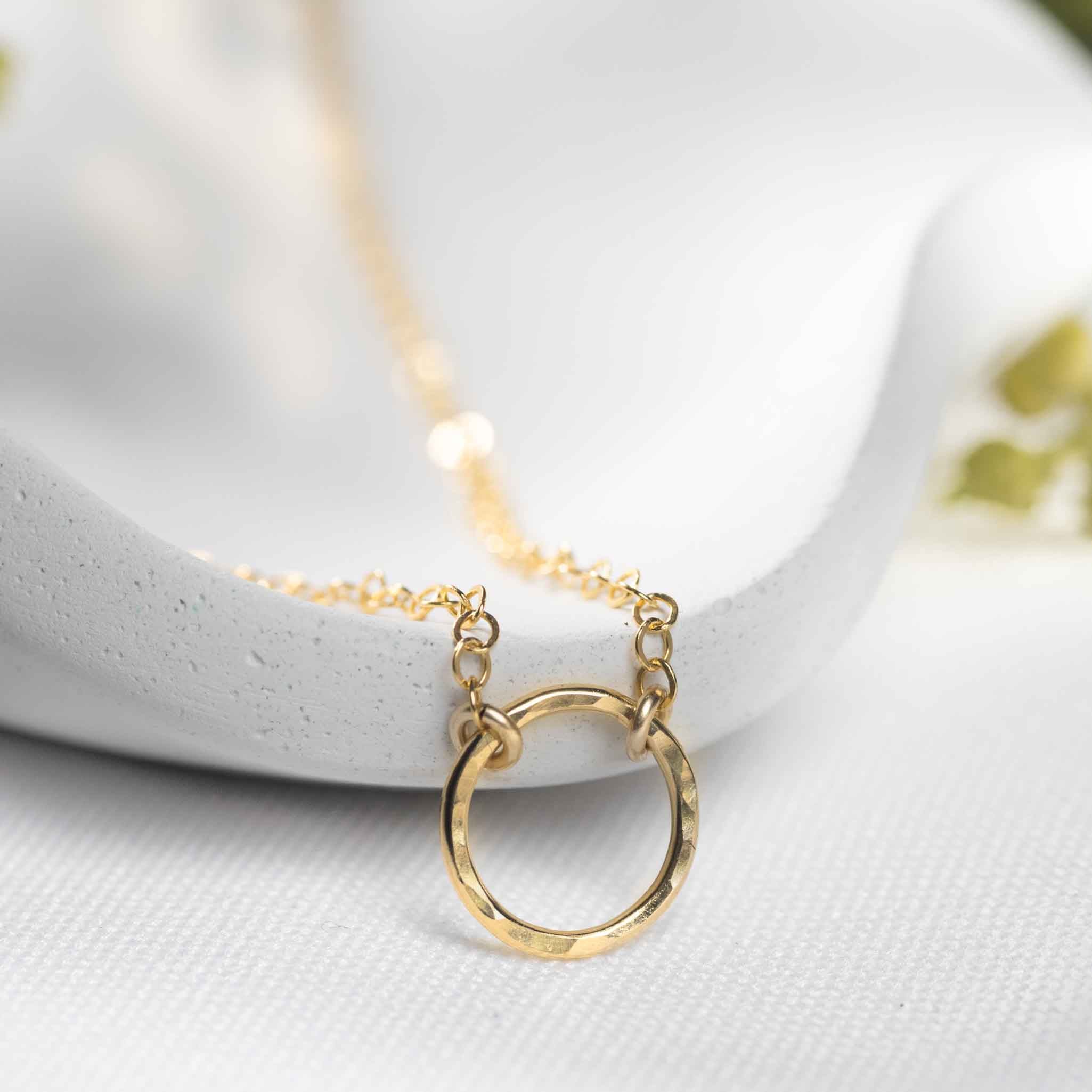Detailansicht des gehämmerten Ringes im Zentrum einer feinen, goldenen Kette, angelehnt an ein weißes Schälchen.