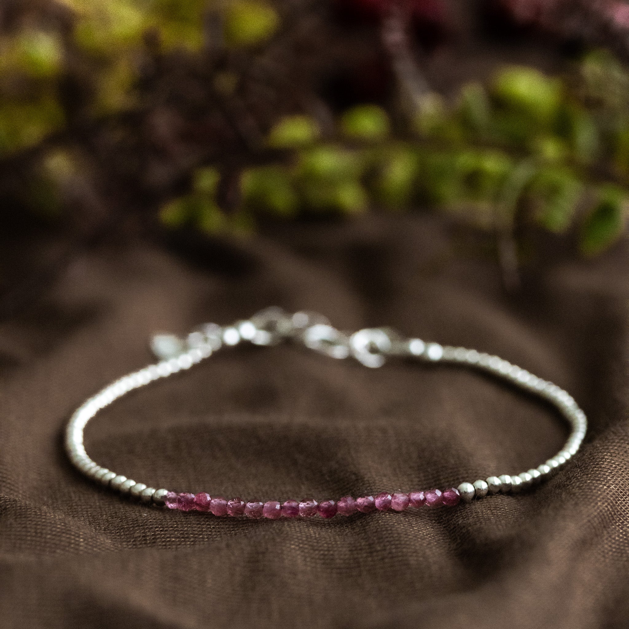 Ein sehr dünnes Silberarmband mit winzigen rosa Turmalinen in der Mitte liegt auf braunem Stoff. Verschwommener floraler Hintergrund.