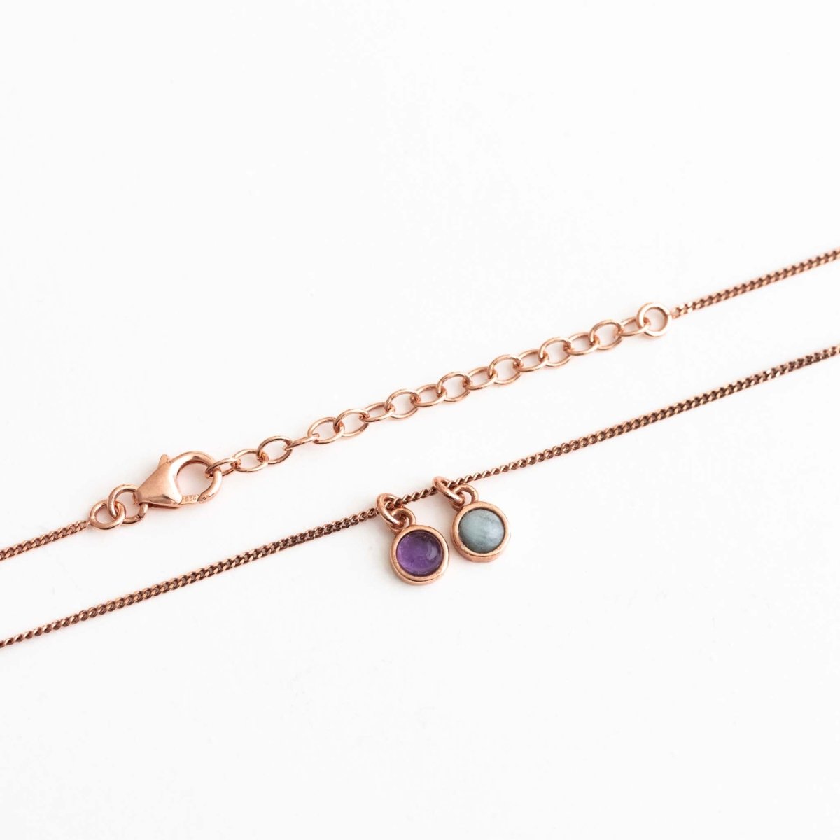 Auf weißem Untergrund liegt eine feine, rosé goldene Panzerkette mit zwei runden Edelstein-Anhängern, einem Amethyst und einem Aquamarin.