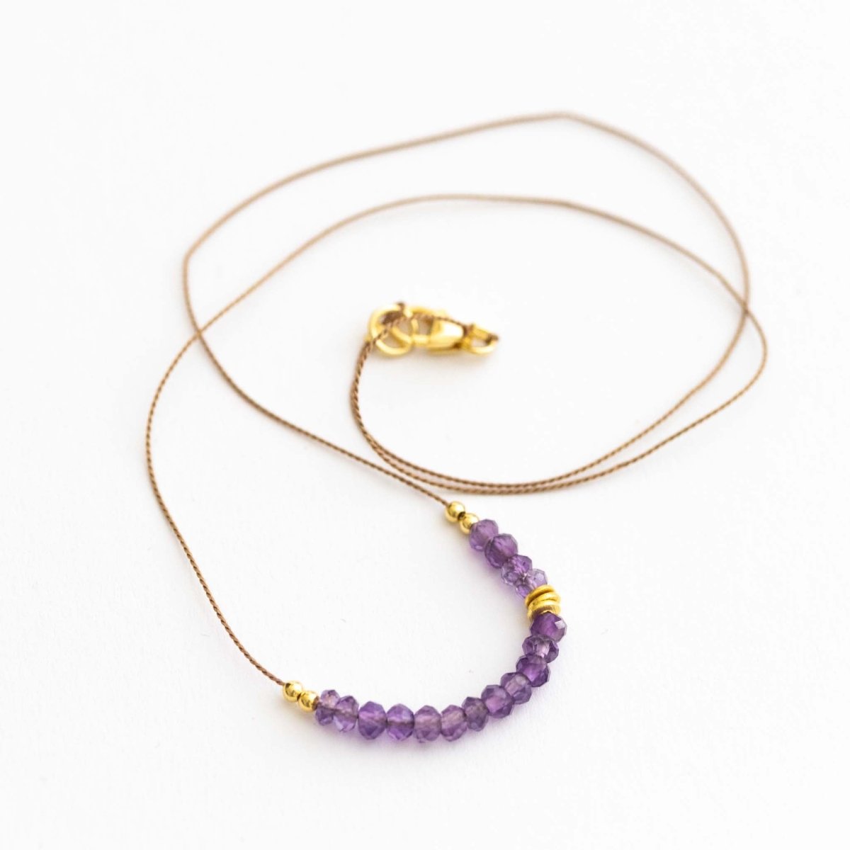 Auf weißen Hintergrund liegt eine kurze Halskette. Sie besteht aus einer dünnen Seidenschnur mit einer Reihe kleiner, violetter Amethyst-Rondellen und goldenen Zierperlen in der Mitte.