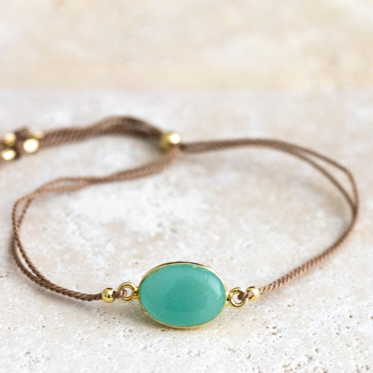 Ein beiges Seidenarmband mit einem gold gefassten, ovalen Aqua Chalcedon liegt auf einer hellen Natursteinplatte.