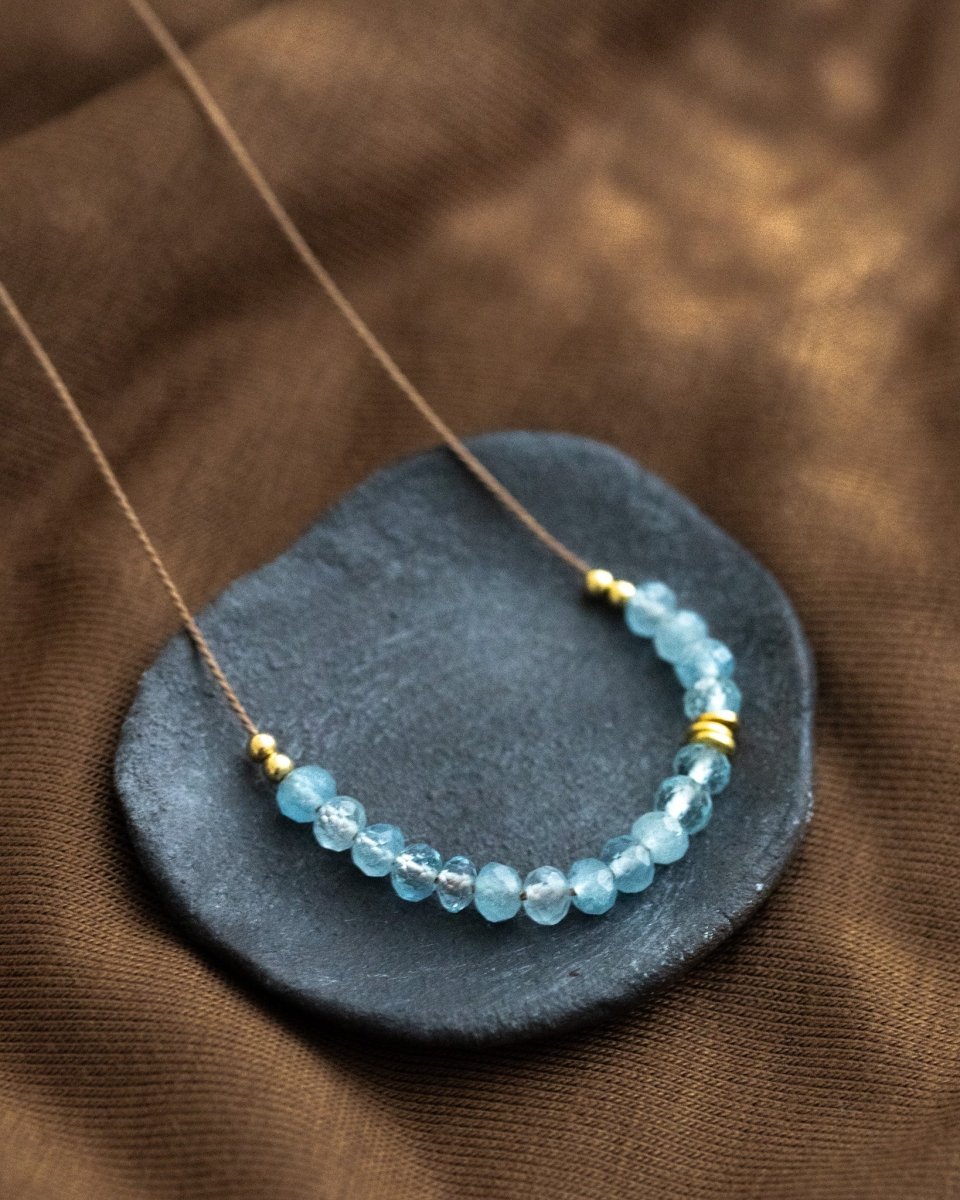 Auf einem dunkelgrauen Schälchen liegt eine Halskette, die aus dünner, beiger Seidenschnur besteht, auf die in der Mitte eine Reihe kleiner Aquamarin-Rondelle gezogen wurden.