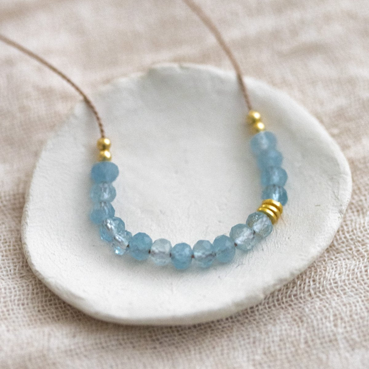 Auf einem weißen Schälchen liegt eine Halskette, die aus dünner, beiger Seidenschnur besteht, auf die in der Mitte eine Reihe kleiner Aquamarin-Rondelle gezogen wurden.
