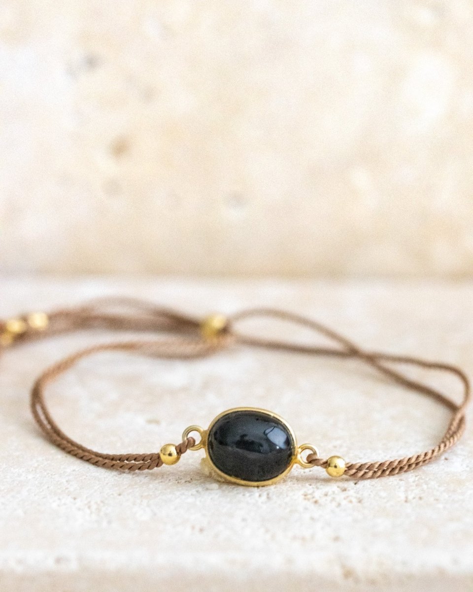 Ein beiges Seidenarmband mit einem Black Star Edelstein in der Mitte auf einer hellen Steinplatte.