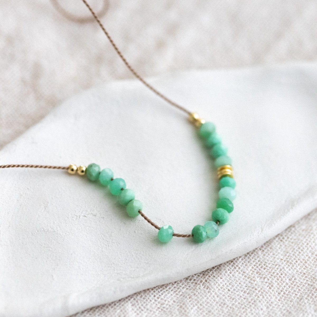 Auf weißem Hintergrund liegt eine dünne Halskette. Sie besteht aus einer beigen Seidenschnur, auf die eine Reihe grüner Chrysopras-Rondelle und einige goldene Zierperlen aufgezogen sind.