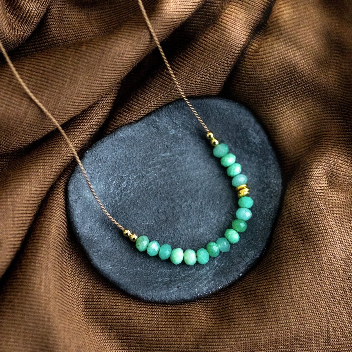 Eine beige Seidenkette mit aufgezogenen, hellgrünen Chrysopras-Steinchen und goldenen Akzenten liegt auf einem grauen Schälchen auf braunem Stoff-Untergrund.