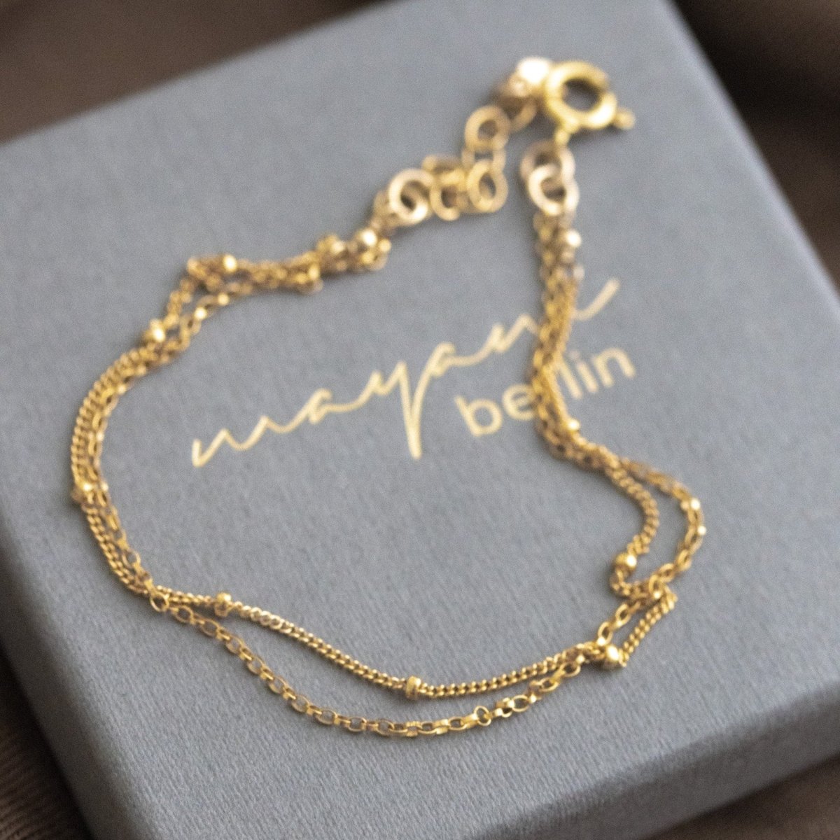 Auf einem grauen Schmucketui liegt ein zierliches, doppelt gelegtes Armband aus Satellitenkette und Rolokette, beide in gold.