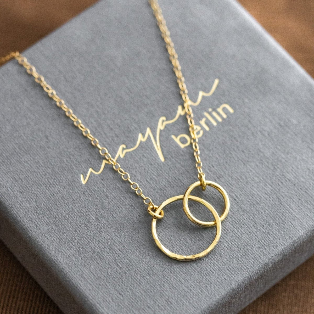 Eine feine Goldkette mit zwei ineinander verschlungenen Ringen liegt auf einem grauen Schmucketui mit der eingeprägten Beschriftung mayam berlin.