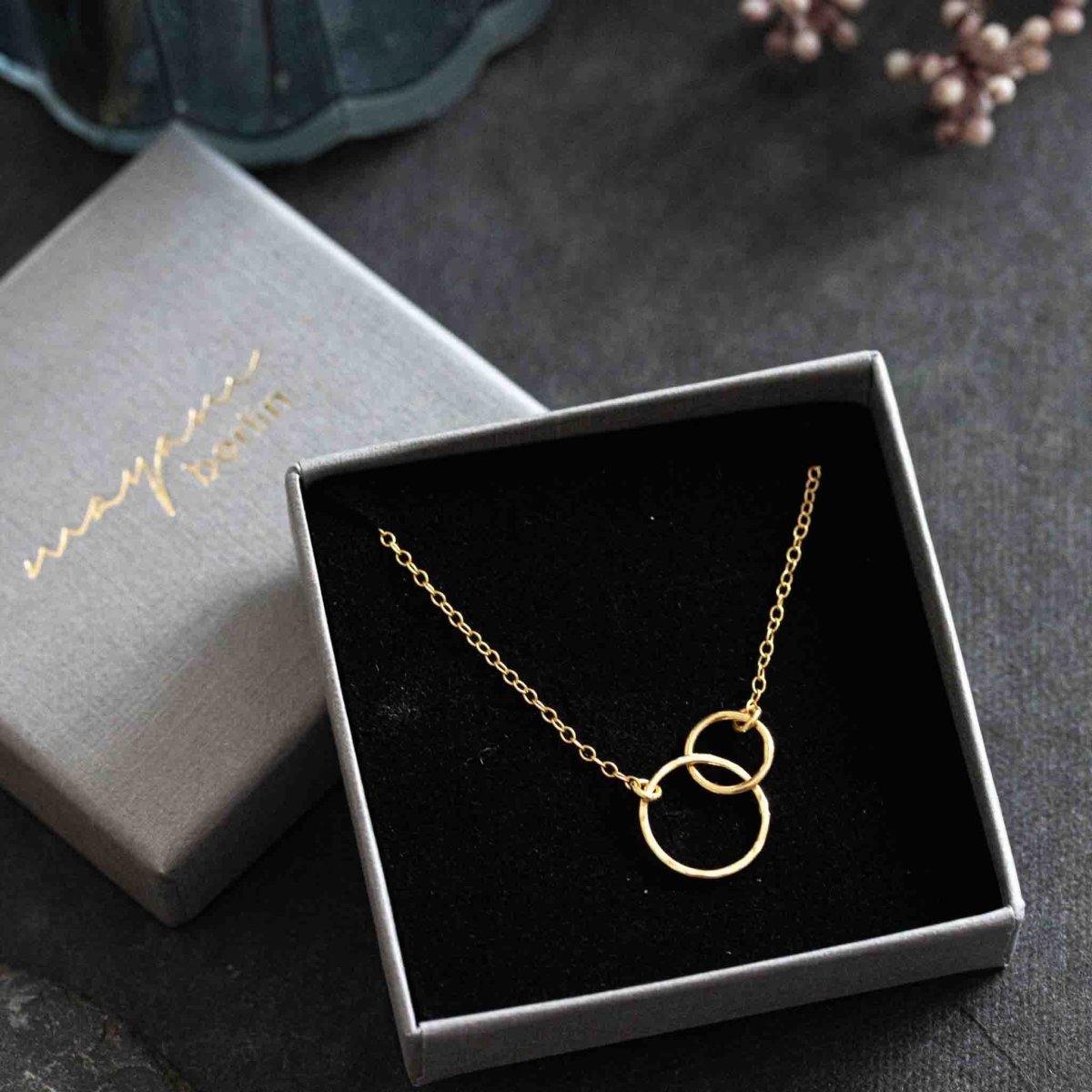 Eine feine Goldkette mit zwei ineinander verschlungenen Ringen ist in den schwarzen Schaumstoff eines grauen Schmucketuis eingehängt.