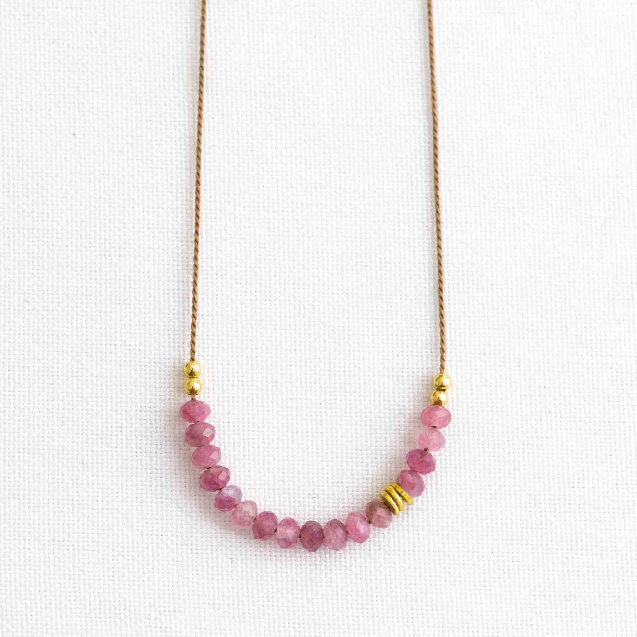 Auf weißen Hintergrund liegt eine kurze Halskette. Sie besteht aus einer dünnen Seidenschnur mit einer Reihe kleiner, rosa Turmalin-Rondelle und goldenen Akzenten in der Mitte.