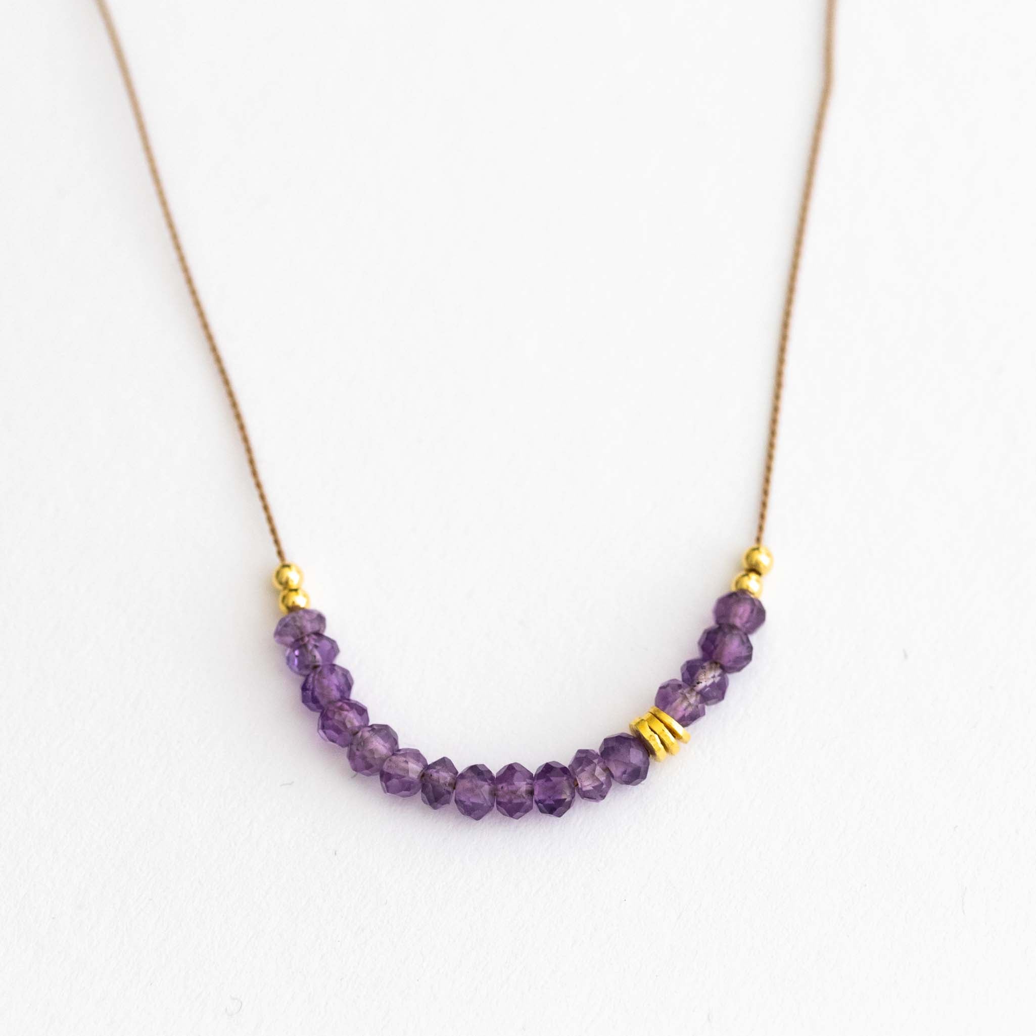 Auf weißen Hintergrund liegt eine kurze Halskette. Sie besteht aus einer dünnen Seidenschnur mit einer Reihe kleiner, violetter Amethyst-Rondellen und goldenen Zierperlen in der Mitte.