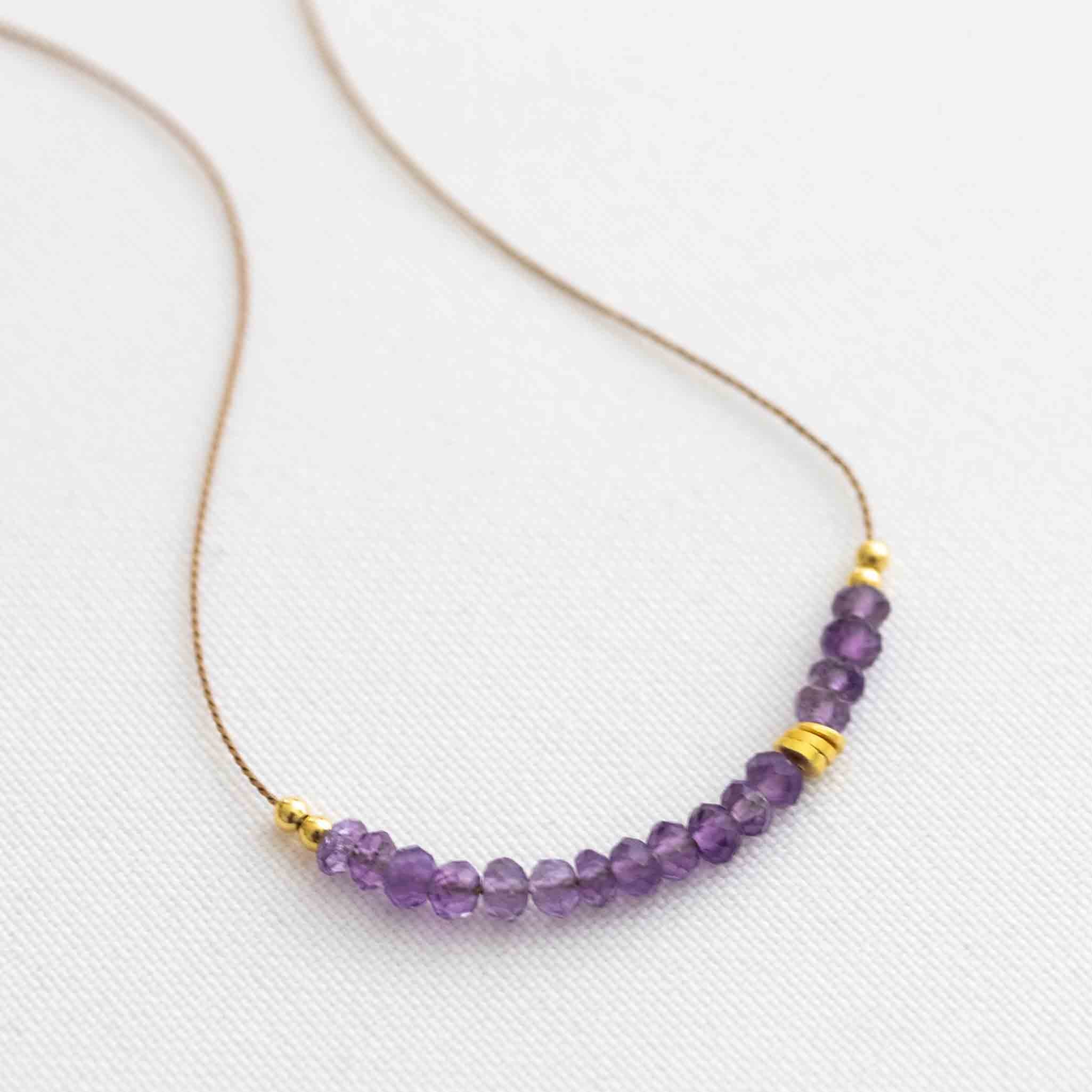 Auf weißen Hintergrund liegt eine kurze Halskette. Sie besteht aus einer dünnen Seidenschnur mit einer Reihe kleiner, violetter Amethyst-Rondellen und goldenen Zierperlen in der Mitte.