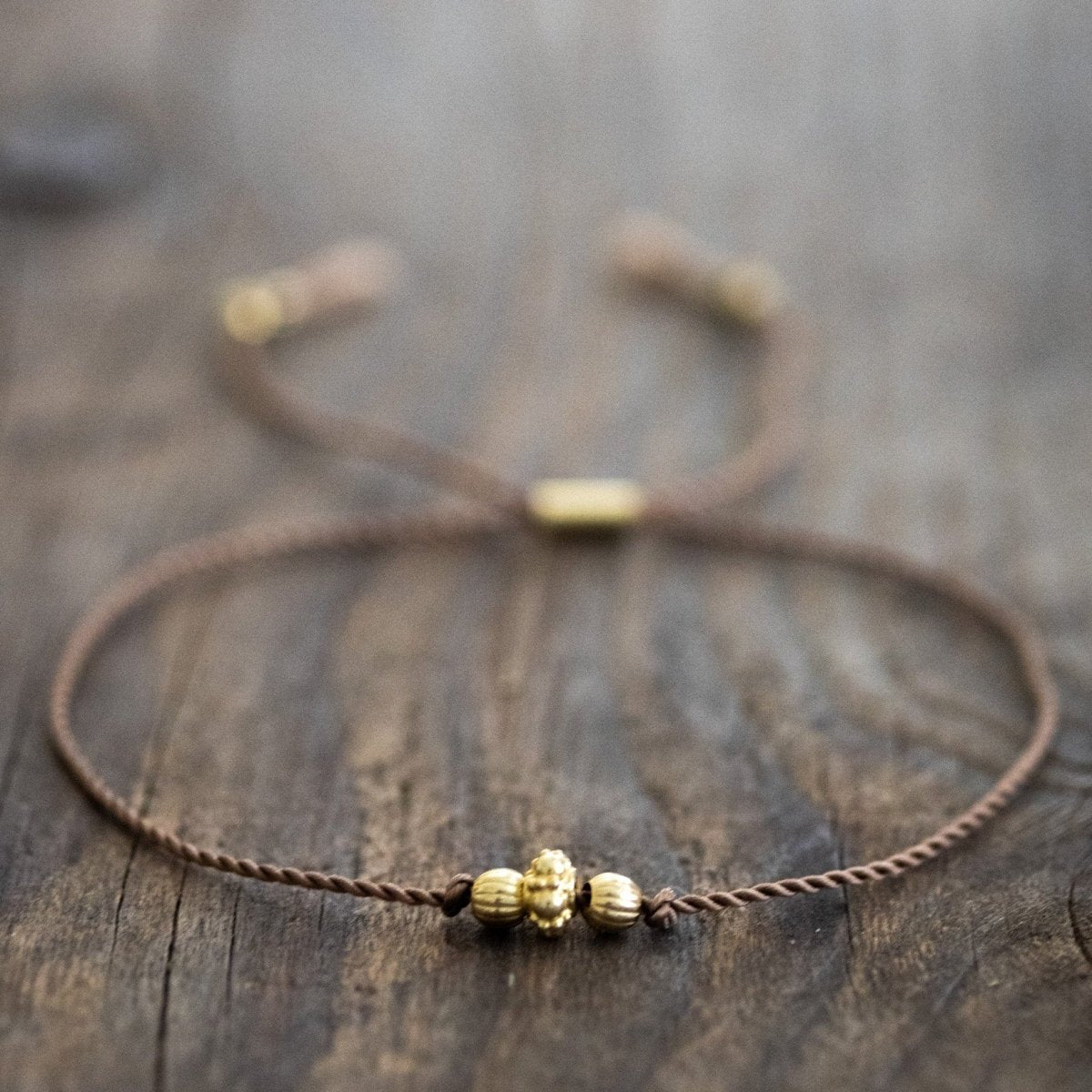 Ein dünnes, braunes Schnurarmband mit drei unterschiedlichen, goldenen Zierperlen in der Mitte auf Holz ausgelegt.
