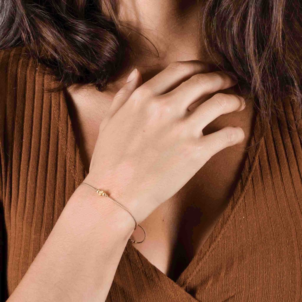 Ein dünnes, braunes Schnurarmband mit drei unterschiedlichen, goldenen Zierperlen in der Mitte am Handgelenk getragen.
