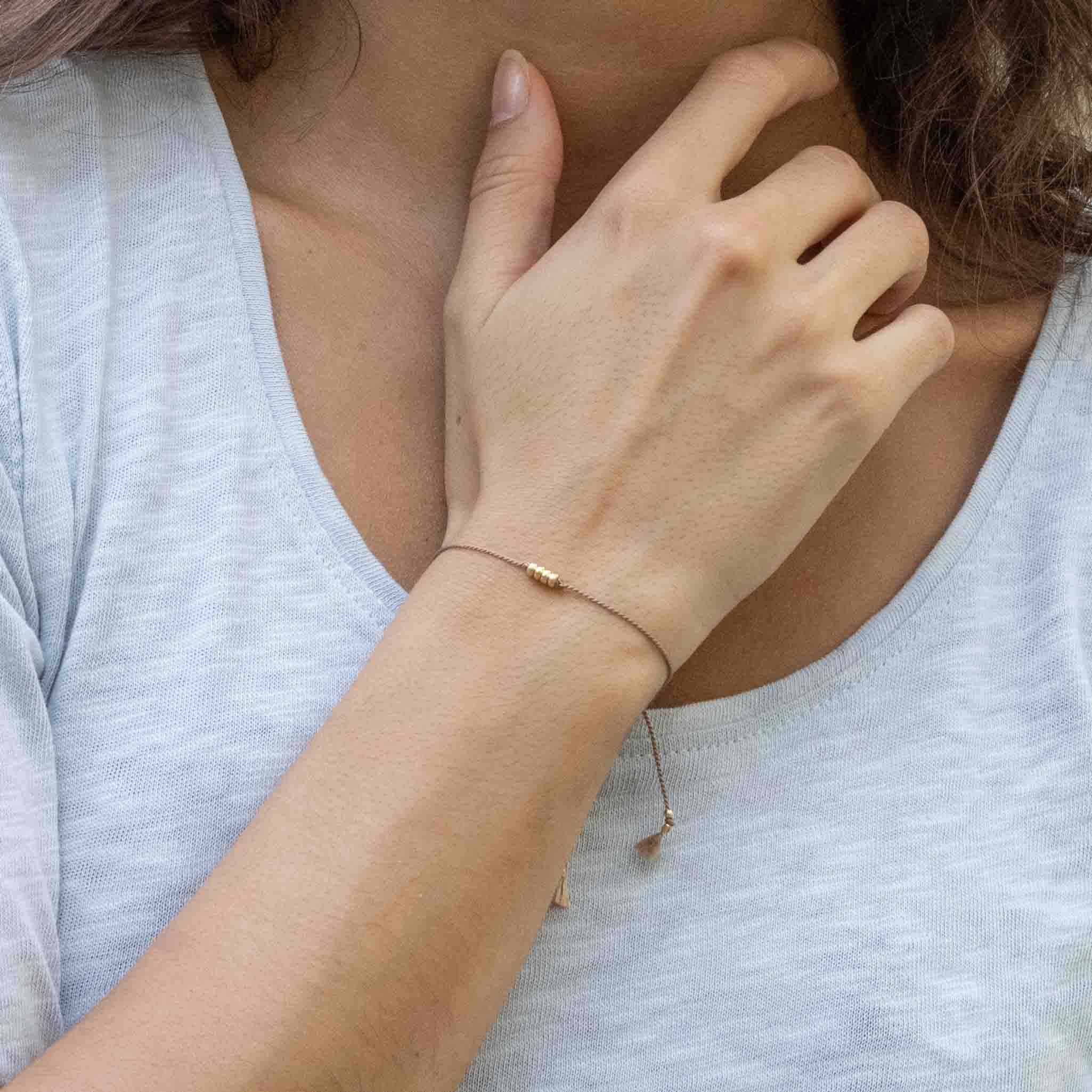 Am Handgelenk des Models ist ein feines Armband zu sehen. Es besteht aus einer beigen Seidenschnur mit vier goldenen Perlen in der Mitte.