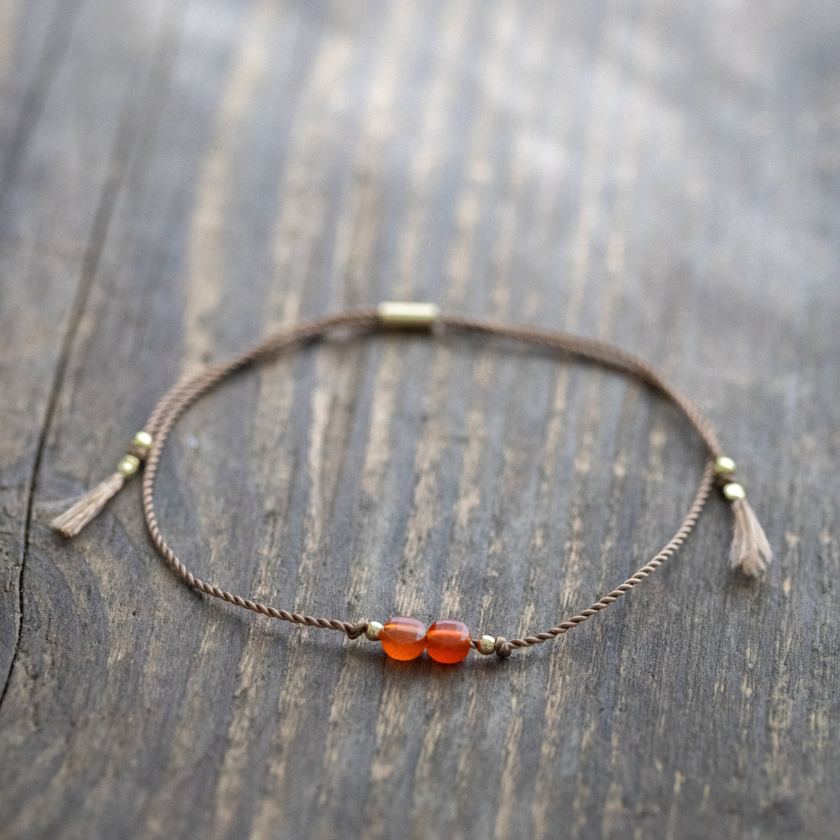 Dünnes Schnurarmband mit orangem Karneol und goldenen Akzenten auf Holz ausgelegt.
