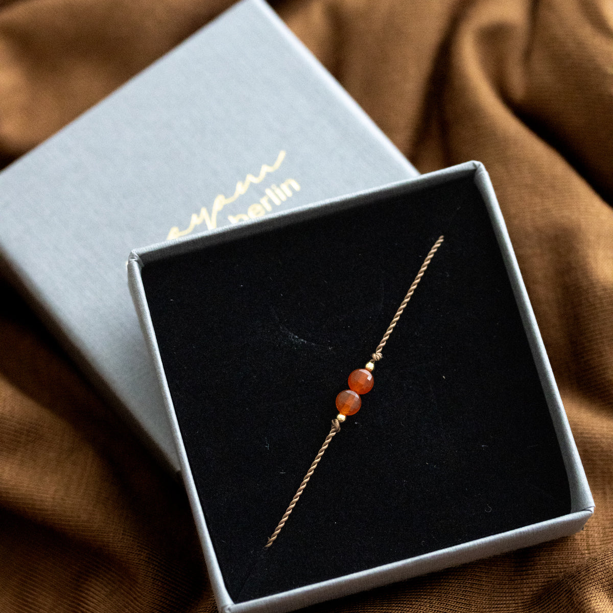 Dünnes Schnurarmband mit orangem Karneol und goldenen Akzenten in den Schaumstoff eines geöffneten Etuis eingehängt.