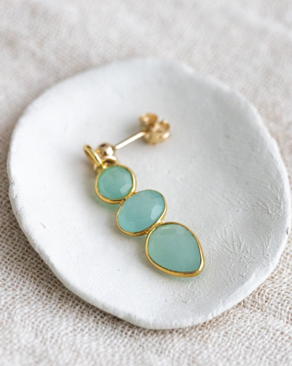 Ein langer, goldener Ohrring mit drei meeresfarbenen Aqua Chalcedonen liegt in einem weißen Schälchen auf weißem Leinenstoff.