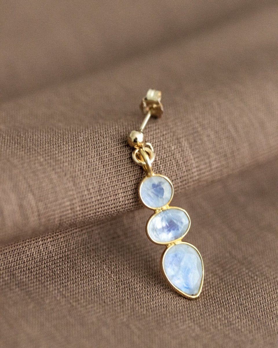 Ein goldener Ohrstecker mit einem dreifachen Mondstein-Anhänger lehnt an braunem Stoff.