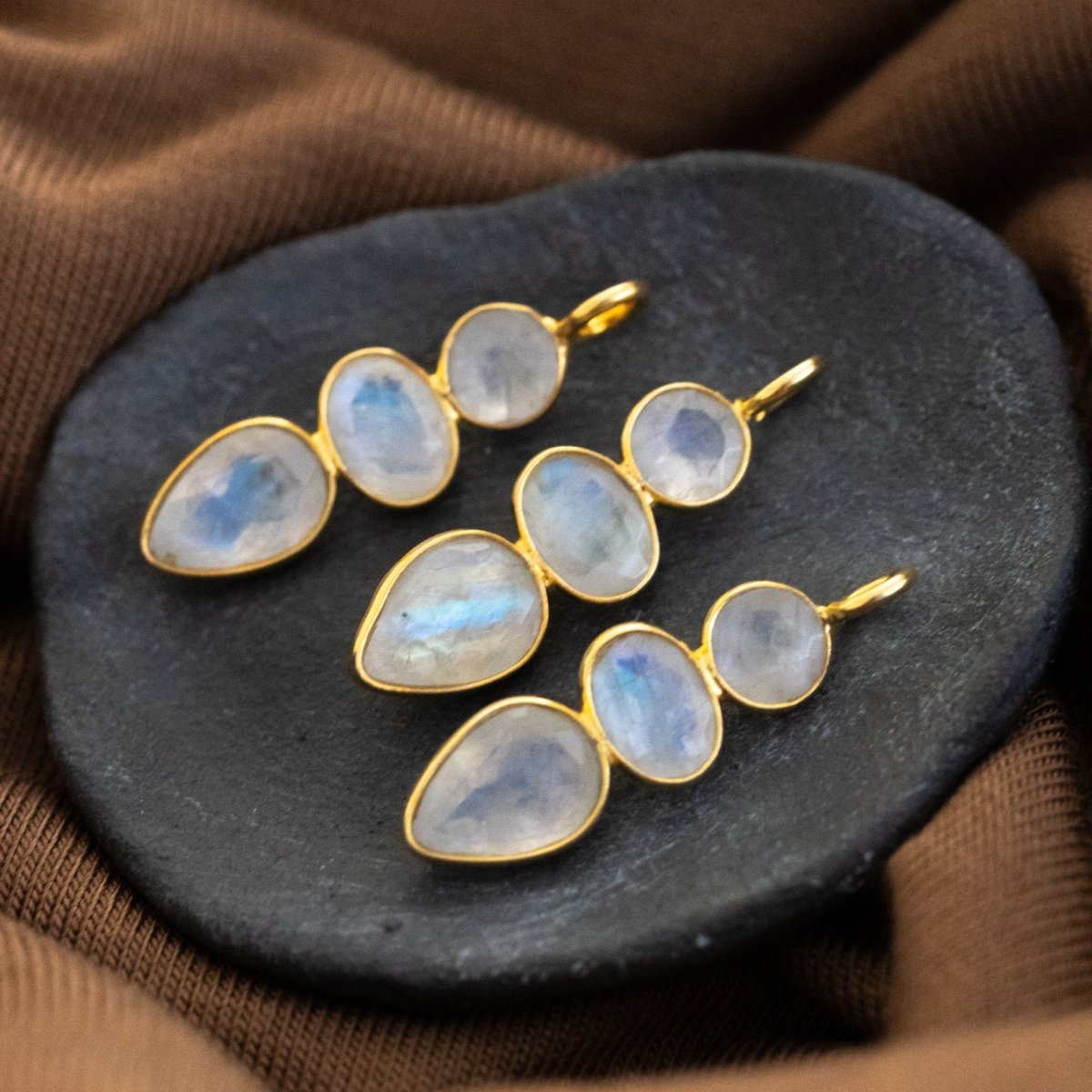 Drei Mondsteinanhänger, die aus jeweils drei miteinander verbundenen Mondsteinen bestehen, liegen auf einem dunkelgrauen Steinschälchen.