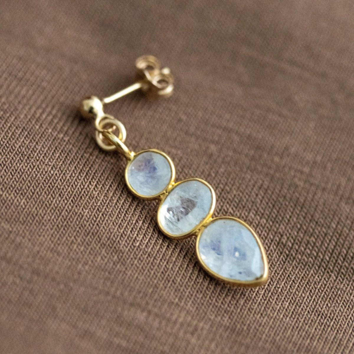 Ein goldener Ohrstecker mit einem dreifachen Mondstein-Anhänger liegt auf braunem Stoff.