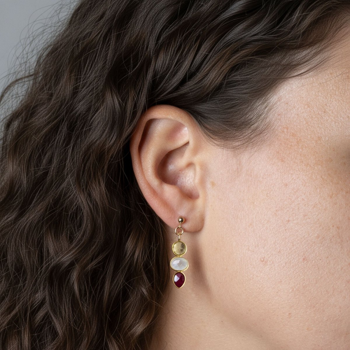 Am Ohr des Models hängt ein länglicher Ohrstecker mit drei gold gefassten Edelsteinen: Citrin, Mondstein und Rubin. Die Steine sind untereinander angebracht.