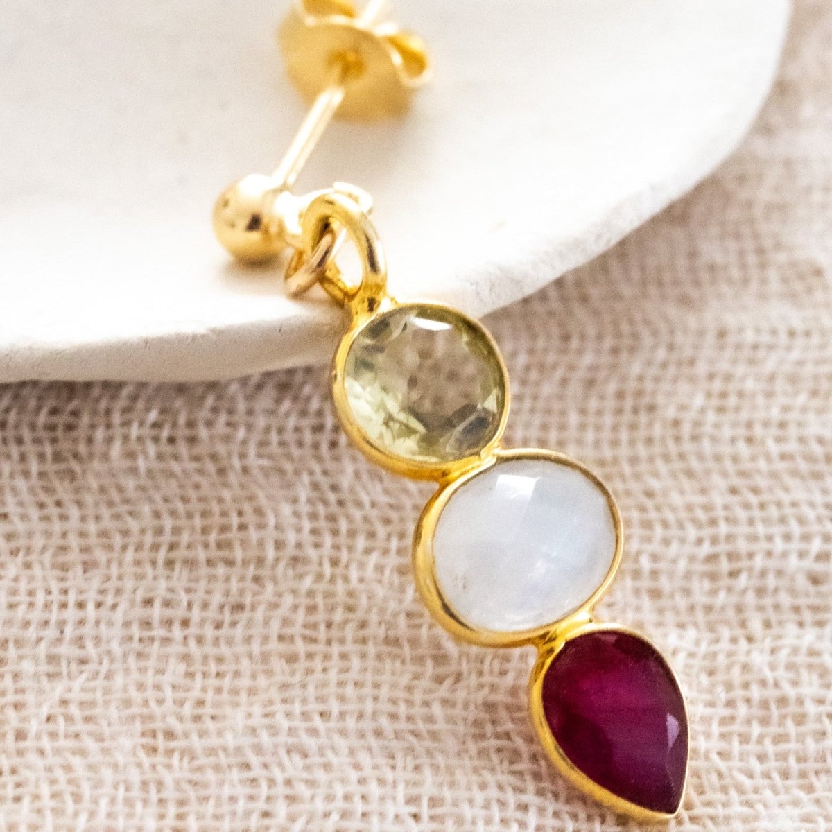 Ein Ohrstecker, bestehend aus Citrin, Mondstein und Rubin, jeweils in gold gefasst, lehnt an einem weißen Schälchen.