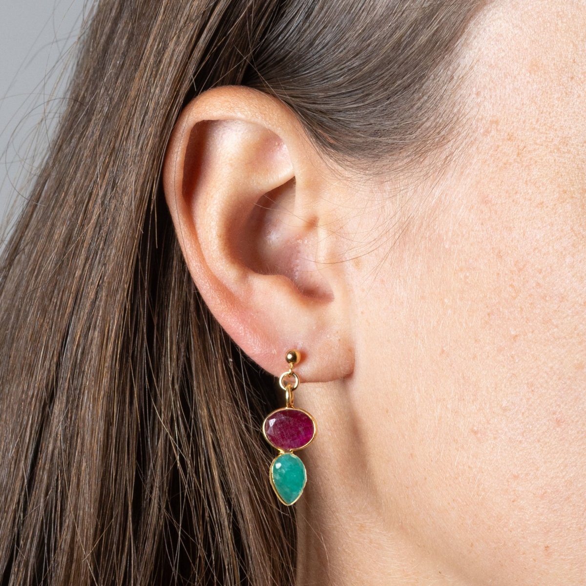 Am Ohr des Models hängt ein goldener Ohrstecker, der aus einem Rubin und einem darunter angebrachten Smaragd besteht. Beide Steine sind gold gefasst.