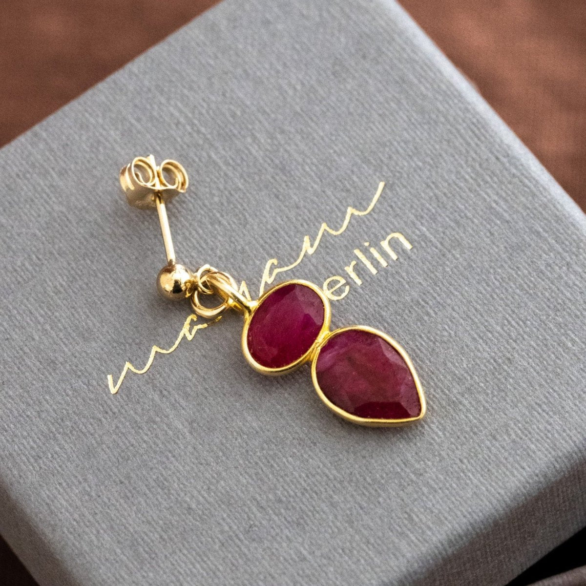 Ein goldener Ohrstecker mit zwei übereinander angebrachten Rubinen liegt auf einem grauen Schmucketui.