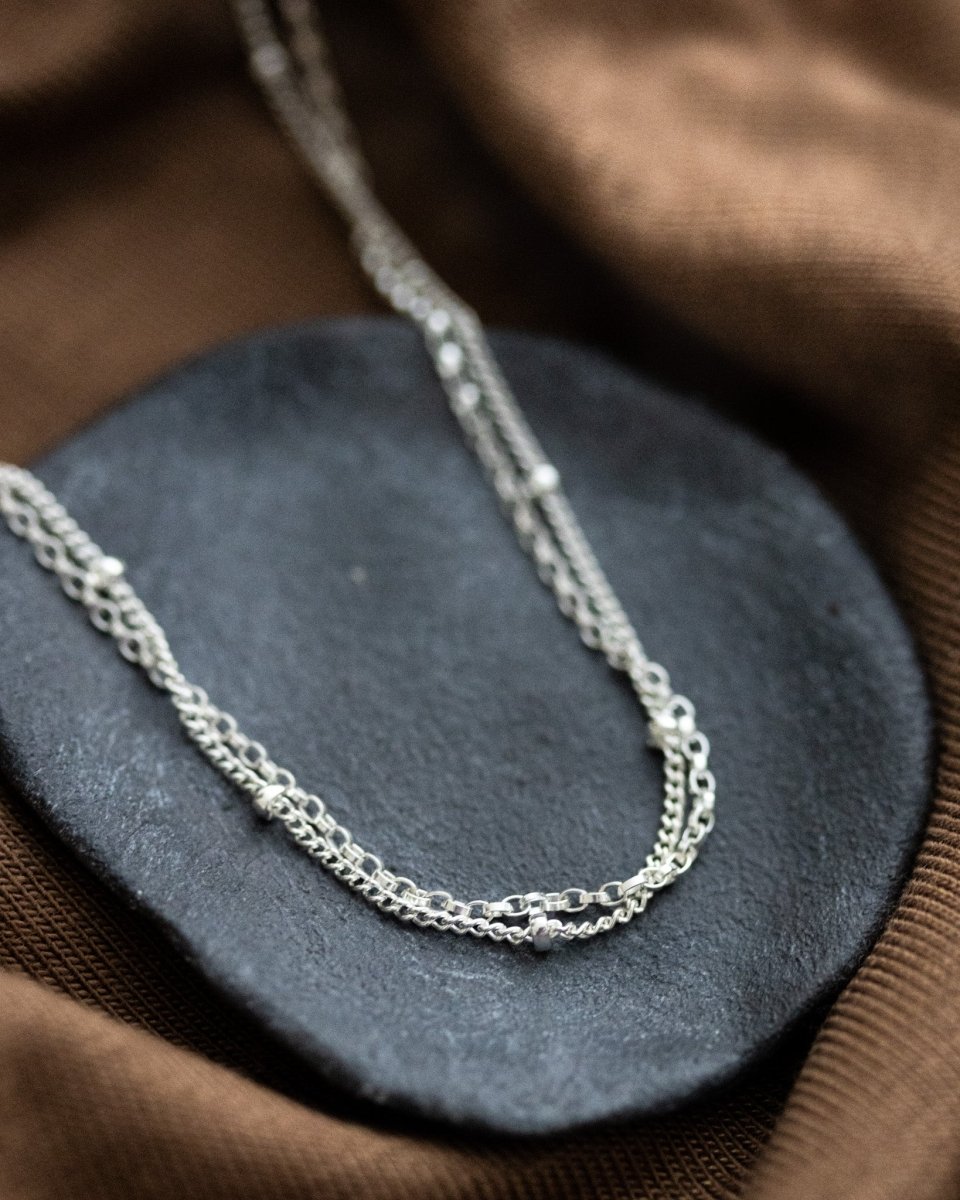 Eine doppelreihige Silberkette, bestehend aus einer Erbskette und einer Satellitenkette, liegt auf einem grauen Steinschälchen, das auf gewelltem, braunen Stoff steht.