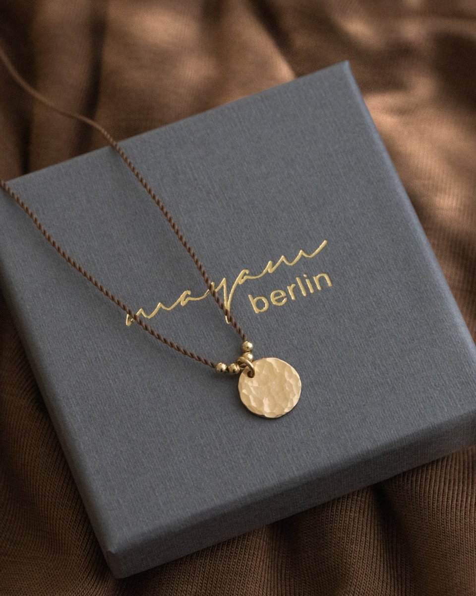 Eine braune Schnurkette mit einem gehämmerten, runden Plättchenanhänger in gold liegt auf einem grauen Schmucketui.