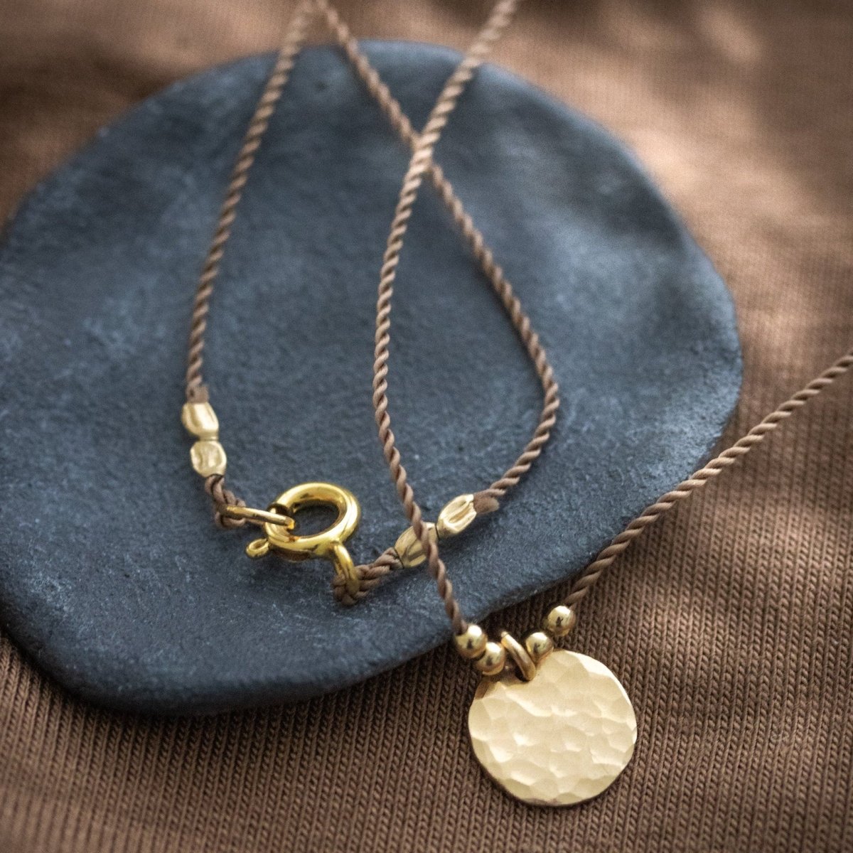 Eine braune Schnurkette mit einem gehämmerten, runden Plättchenanhänger in gold liegt auf einem schwarzen Steinschälchen.