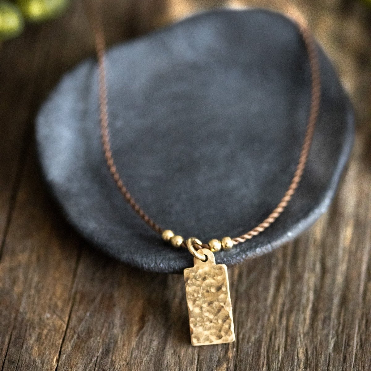 Eine feine, beige Kordelkette mit einem rechteckigen, gehämmerten Plättchenanhänger lehnt an einem dunkelgrauen Steinschälchen auf braunem Holzuntergrund.