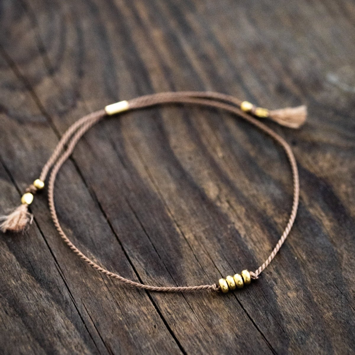 Ein dünnes, beiges Schnurarmband mit vier Goldperlen in der Mitte liegt auf braunem Holz.
