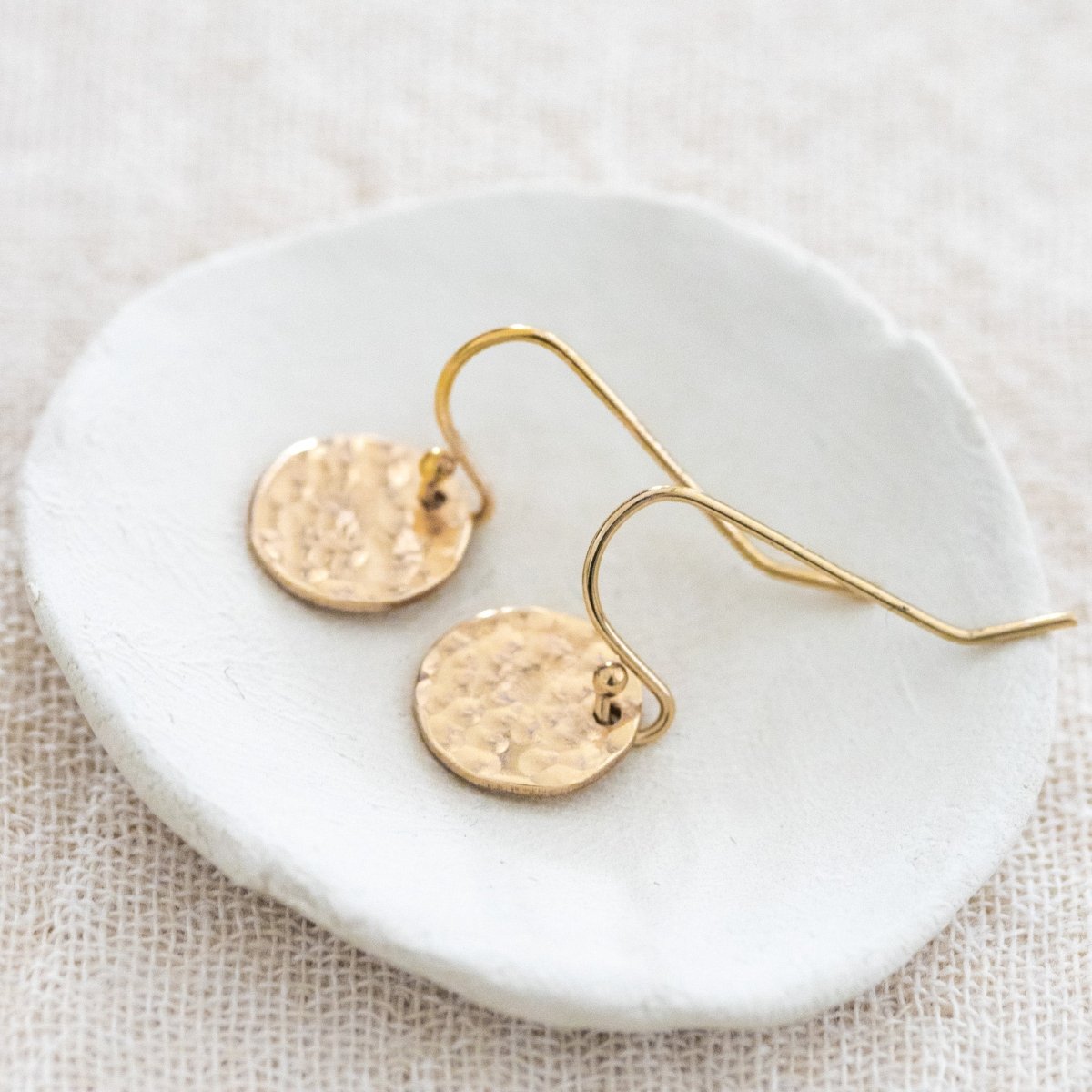 Ein Paar Gold-Ohrringe mit runden, gehämmerten Plättchen an Ohrhaken liegt auf einem weißen Schälchen.