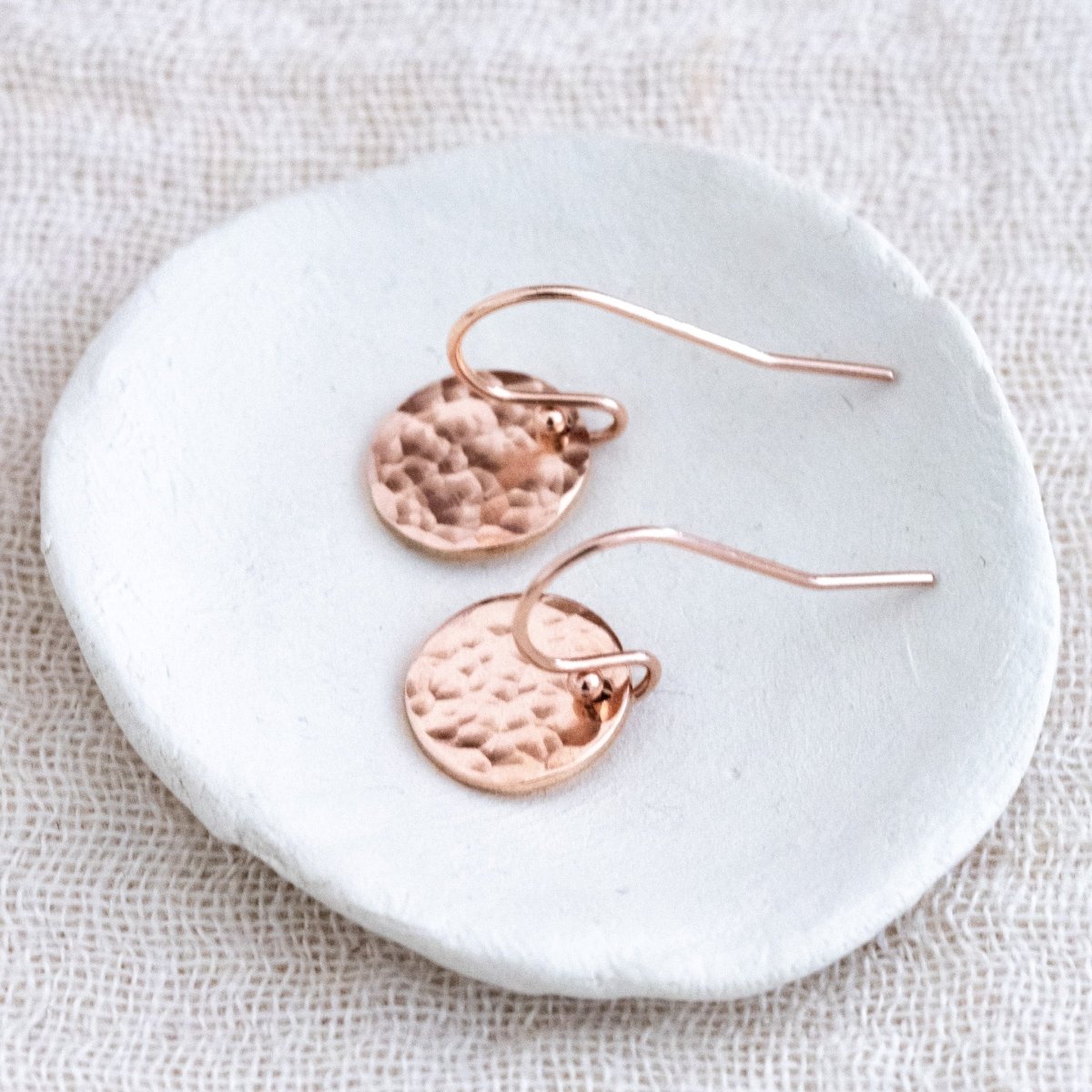 Ein Paar rosé goldener Ohrringe mit je einem runden, gehämmerten Plättchen an Ohrhaken  liegt auf einem weißen Schälchen.