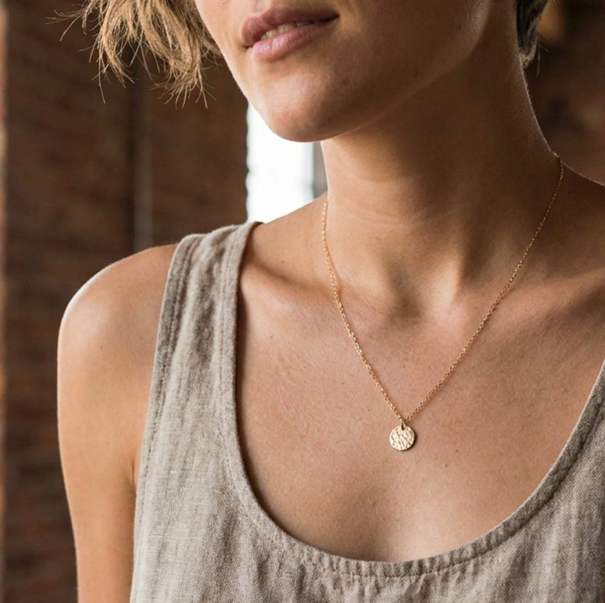 Eine feine und kurze Goldkette mit einem runden, gehämmerten Plättchen-Anhänger hängt vom Hals des Models herab. 