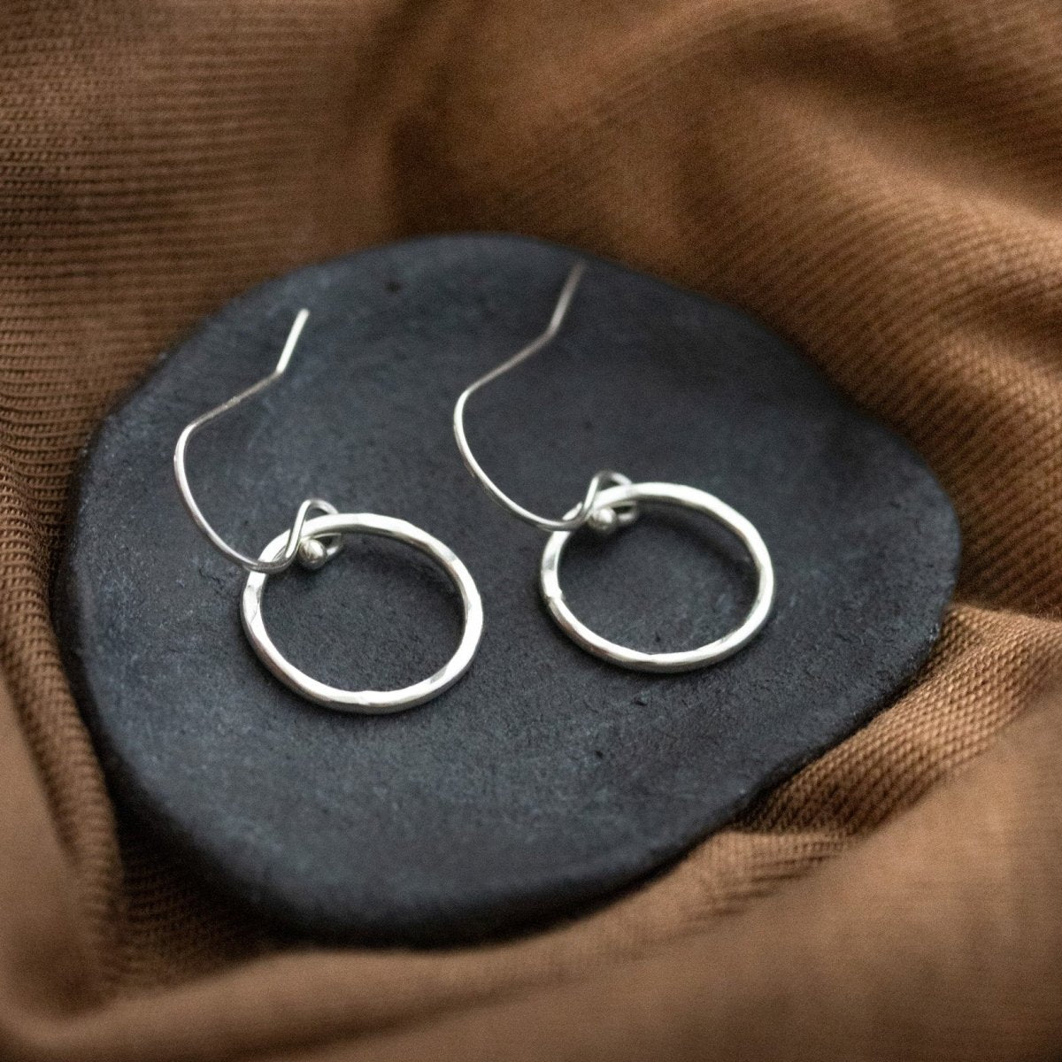 Ein Paar Silber-Ohrringe, bestehend aus jeweils einem gehämmerten Ring an Ohrhaken, liegt auf einem grauen Steinschälchen, das auf braunem Stoff liegt.