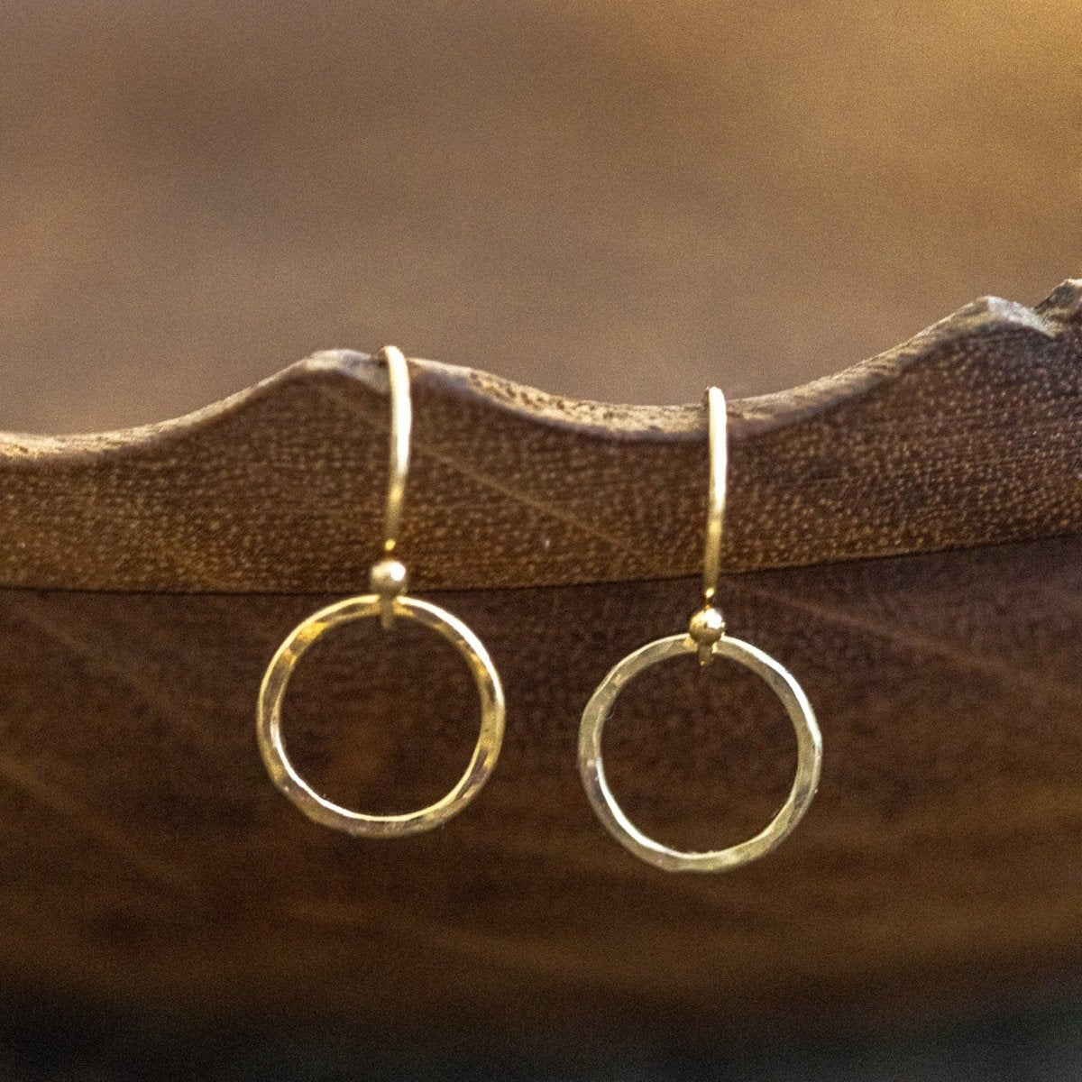 Ein Paar Silber-Ohrringe, bestehend aus jeweils einem gehämmerten Ring an Ohrhaken, hängt an einer braunen Holzschale vor unscharfem Hintergrund.