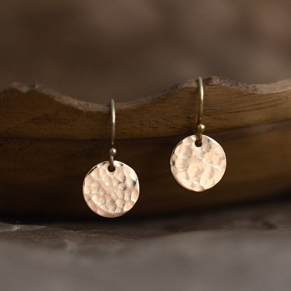 Ein Paar goldener Ohrringe hängen von einer braunen, gewellten Schüssel herab. Die Ohrringe bestehen aus einfachen Ohrhaken, an denen jeweils ein rundes, gehämmertes Plättchen hängt.