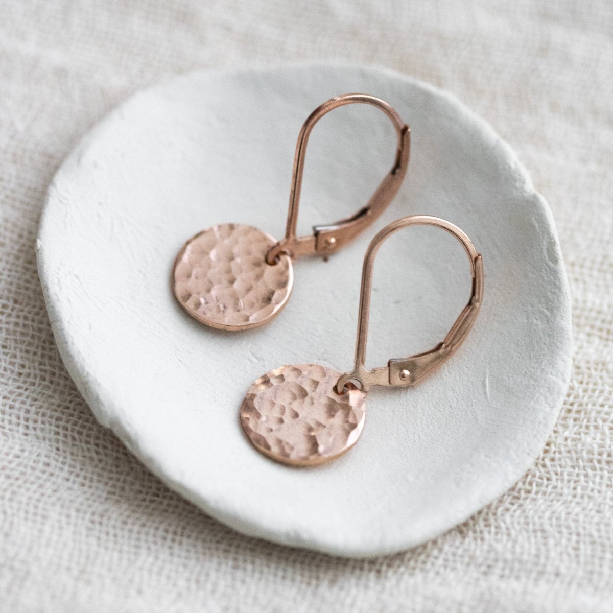 Ein Paar rosé goldener Ohrringe mit je einem runden, gehämmerten Plättchen an einer Klappbrisur liegt auf einem weißen Schälchen.