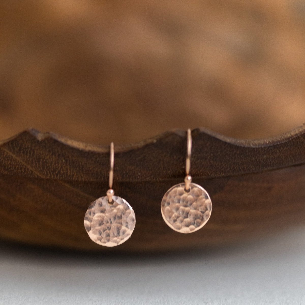 Ein Paar rose goldener Plättchen-Ohrringe mit gehämmerter Oberfläche hängt von einer gewellten, dunklen Holzschüssel herab.