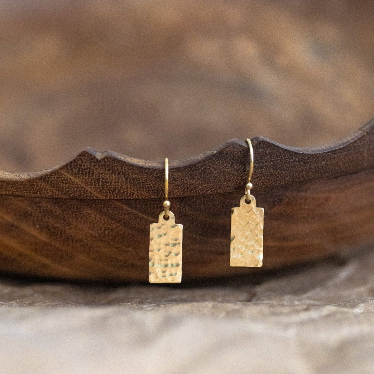 Ein Paar goldener Ohrringe, bestehend aus Klappbrisuren und gehämmerten Plättchen hängt von einem gewellten Holzschälchen herab.