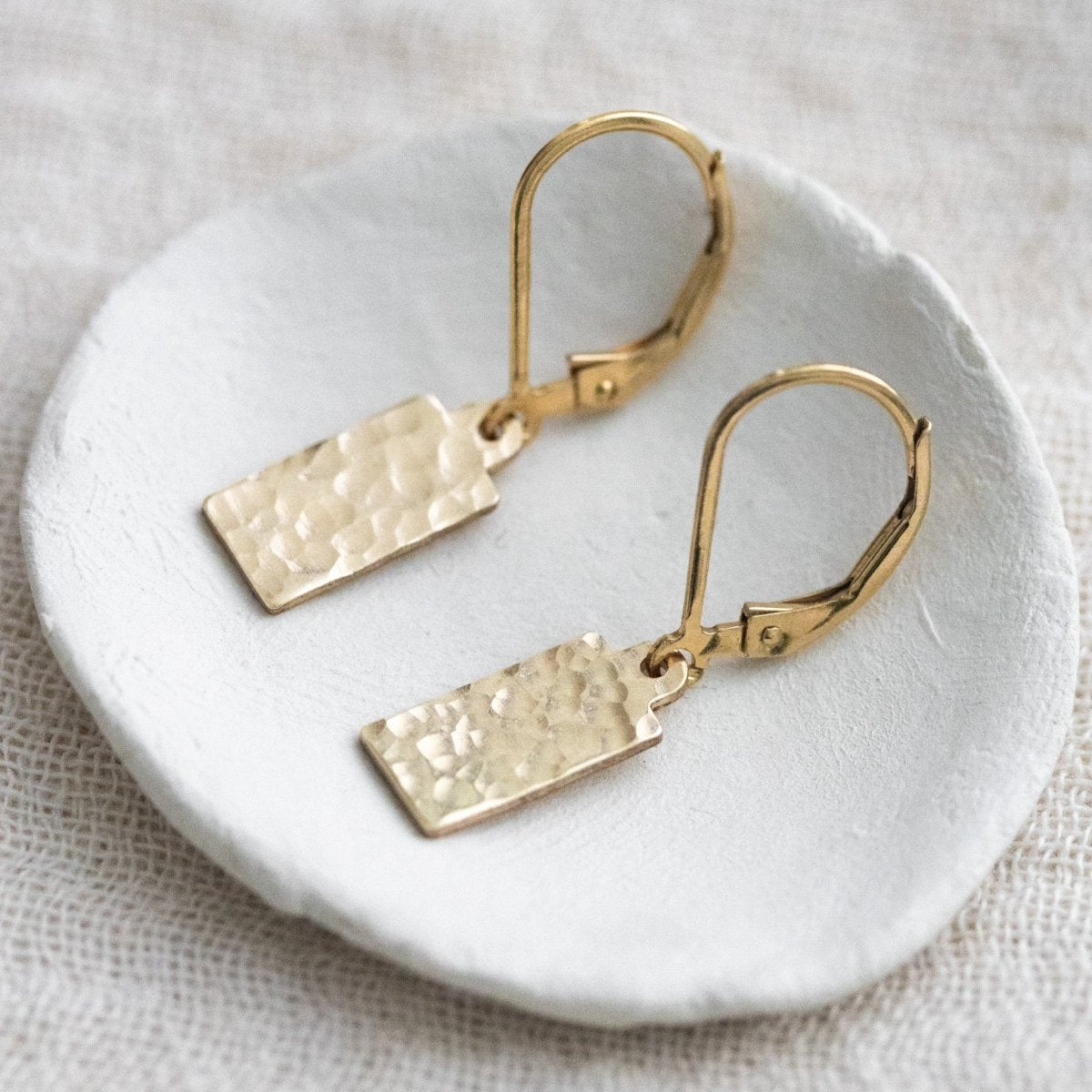 Auf einem weißen Schälchen liegt ein Paar goldener Ohrringe mit jeweils einem rechteckigen Plättchen an einer Klappbrisur.
