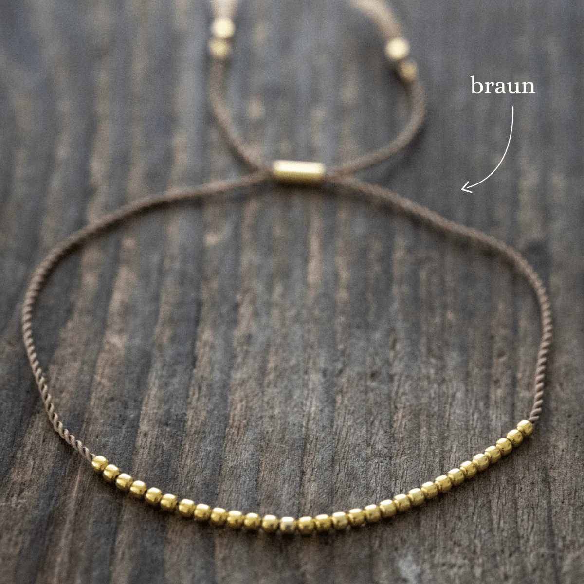 Ein dünnes, braunes Schnurarmband mit vielen goldenen Perlen und einem Schiebeverschluss liegt auf braunem Holz.