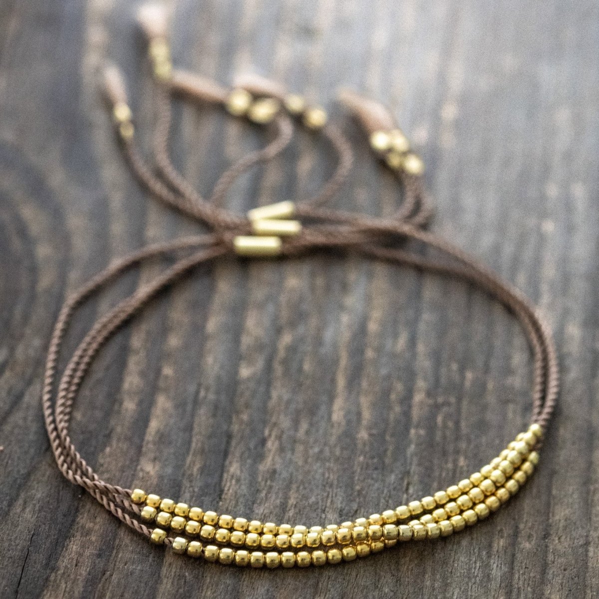 Drei feine, braune Schnurarmbänder mit einer Reihe winziger Goldperlen liegt auf verwittertem Holz.