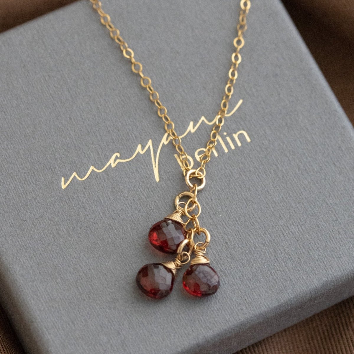 Eine feine Goldkette mit drei untereinander hängenden Granat Edelsteinen liegt auf einem grauen Schmucketui.